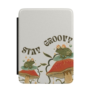 May include: A white tablet case featuring the text STAY GROOVY in dark green lettering. The design includes two green frogs wearing orange flower crowns, each perched atop a red and tan mushroom, surrounded by small stars and bubbles.