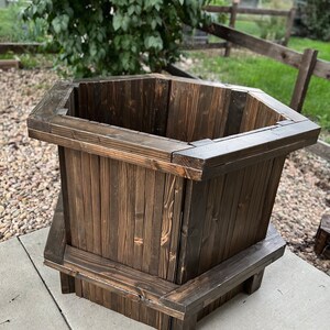 May include: A dark brown hexagonal wooden planter with a wide opening and a slightly flared base. The planter is made of wood planks and has a rustic, weathered look.