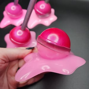 May include: Three pink candy lollipops with white swirls on top, each sitting on a pink, melted-looking base. The lollipops are on a dark background.