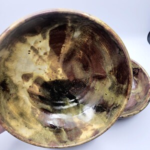 May include: Two ceramic bowls with a mottled glaze in shades of brown, gold, and black. The bowls have a rustic, organic appearance, with visible texture and variations in color. The bowls are likely handmade and suitable for serving food.