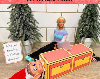 May include: A Christmas-themed scene featuring a doll, an elf, and a magic trick box. The box is yellow and red, with a saw and a sign that reads, "Who said two elves are better than one?!" The text "ELF MAGIC TRICK" is at the top.