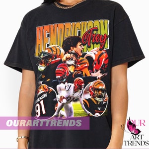 May include: Black t-shirt with a vintage-style graphic of a football player wearing a Cincinnati Bengals jersey. The text "Hendrickson Trey" is printed on the shirt.