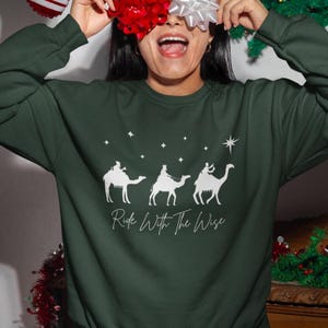 May include: Dark green sweatshirt with a white graphic of three camels and the text "Ride With The Wise". The design includes stars and a bright star. The sweatshirt is a crew neck style.