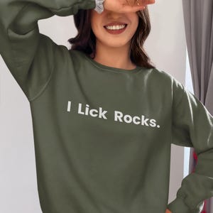 May include: Olive green sweatshirt with the text "I Lick Rocks" in white. The sweatshirt is a solid color and has a crew neck. The text is centered on the front of the shirt.