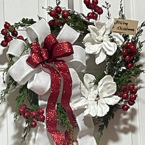 May include: A Christmas wreath with a white bow, red berries, and white magnolia flowers. The wreath has a sign that says "Merry Christmas".