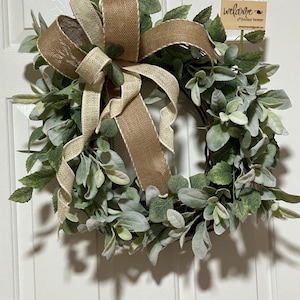 May include: A decorative wreath featuring a mix of green and gray leaves, accented with a large burlap bow. A small wooden sign with the word "welcome" is attached. The wreath is designed for home decor.