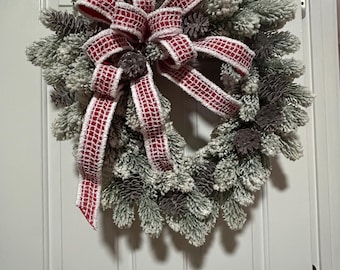May include: A Christmas wreath with frosted pine needles, pine cones, and a red and white plaid ribbon bow. A small wooden sign that says "Merry Christmas" is attached to the wreath.