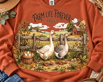 Puede incluir: Una sudadera de cuello redondo color óxido con un estampado gráfico de dos gansos frente a una valla de madera, una cesta de huevos y un granero. El texto "FARM LIFE FOREVER" está impreso en letras marrones sobre la escena.
