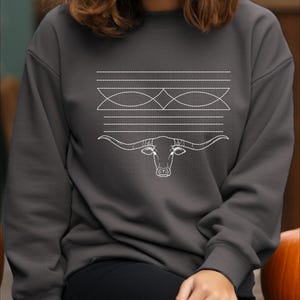 May include: A charcoal gray long-sleeve sweatshirt featuring a white line-art graphic on the chest. The design includes horizontal dashed lines, geometric shapes, and a detailed outline of a longhorn steer head centered below.