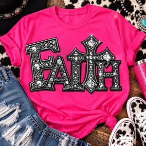 May include: A bright hot pink short-sleeved t-shirt featuring the word FAITH printed in a large, bold, black font. The letters are filled with a decorative pattern of silver rhinestones and studs, with a cross replacing the letter T.