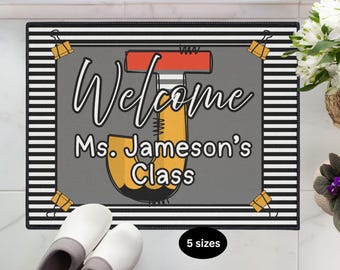 May include: Rectangular welcome mat with a gray background and the words "Welcome Ms. Jameson's Class" in white script. The mat has a black and white striped border and is decorated with yellow binder clips.