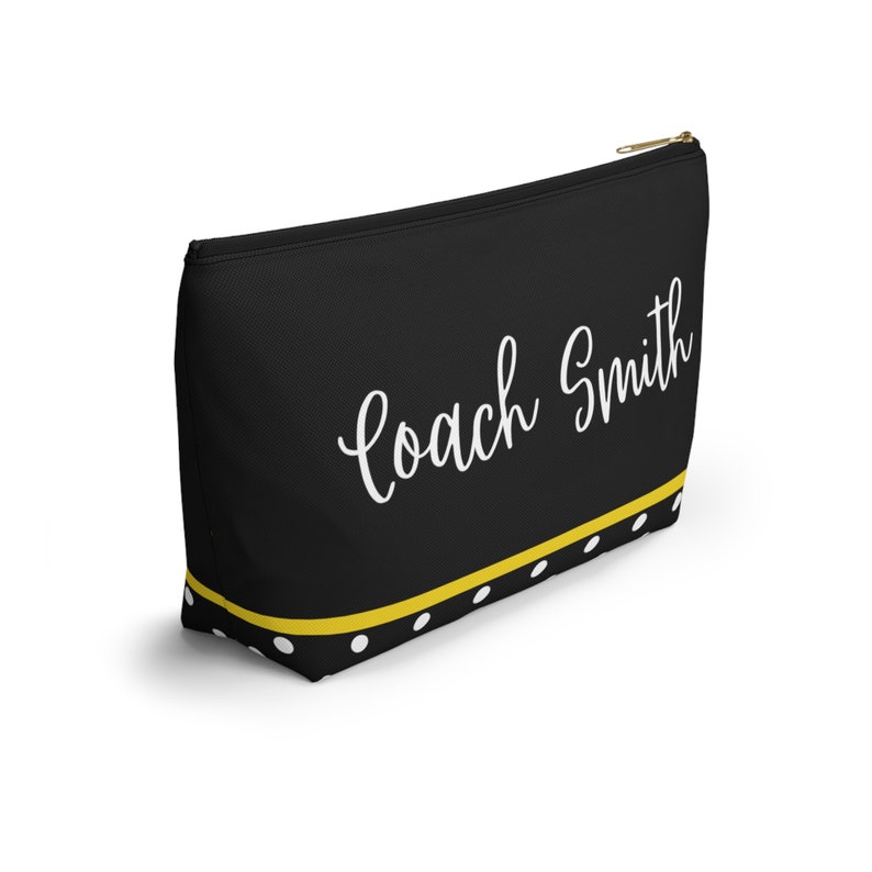 K&ouml;nnte beinhalten: Schwarze Kosmetiktasche mit Rei&szlig;verschluss, gelbem und wei&szlig;em Tupfenrand und dem Text "Coach Smith" in wei&szlig;er Kursivschrift.