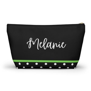 K&ouml;nnte beinhalten: Schwarze Kosmetiktasche mit Rei&szlig;verschluss, gr&uuml;nem Streifen und wei&szlig;en Punkten. Die Tasche tr&auml;gt den Namen "Melanie" in wei&szlig;er Kursivschrift.