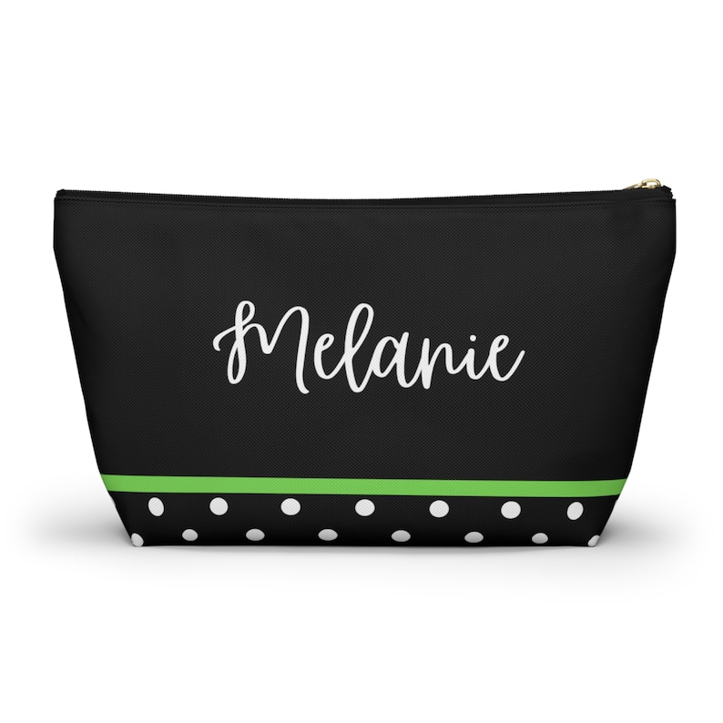 K&ouml;nnte beinhalten: Schwarze Kosmetiktasche mit Rei&szlig;verschluss, gr&uuml;nem Streifen und wei&szlig;en Punkten. Die Tasche tr&auml;gt den Namen "Melanie" in wei&szlig;er Kursivschrift.