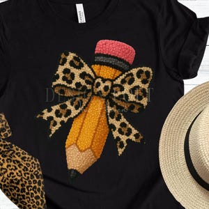 May include: Black t-shirt featuring a large pencil design with a leopard print bow. The pencil is yellow with a pink eraser and black tip. A straw hat is visible in the background.