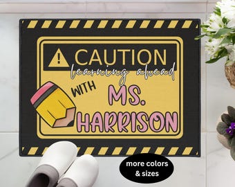 May include: Black rectangular doormat with a yellow caution sign that reads "CAUTION learning ahead with Ms. Harrison." A cartoon pencil graphic is on the left. The border has yellow and black stripes.