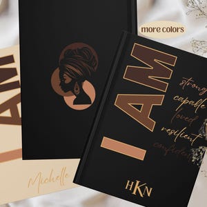 May include: Two black journals with gold accents. One journal features a silhouette of a woman with a headscarf and the words "I AM" in gold. The other journal features the words "I AM" in gold and a list of positive affirmations: strong, capable, loved, resilient, confident.