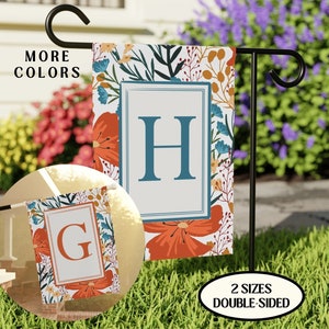 May include: Two double-sided garden flags with floral designs and a large initial letter. The flag on the left has the letter "G" and the flag on the right has the letter "H". The flags are displayed on black metal poles in a grassy area.