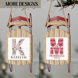 May include: Two wooden Christmas ornaments shaped like sleds with red accents. The ornament on the left has a white background with a red and green patterned letter K and the name Katelyn. The ornament on the right has a white background with a red and white patterned letter M and the text Morrison family.