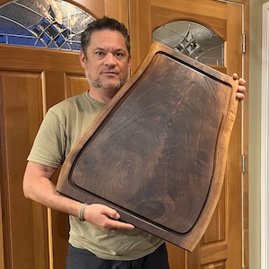 May include: A large, dark brown wooden chopping board with a juice groove and a curved edge. The board is held by a person in front of a wooden door. The wood grain is visible, and the board appears to be hand-crafted.