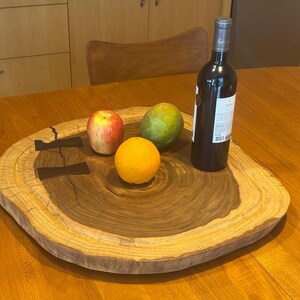 May include: A wooden serving platter with a natural edge, displaying an apple, mango, and orange. A bottle of red wine is placed beside the fruit. The tray has dark brown inlaid details and is on a wooden table.