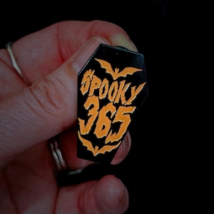 May include: Black coffin-shaped enamel pin with orange text and bat silhouettes. The pin reads "Spooky 365" in a stylized font, perfect for Halloween enthusiasts. A unique accessory for jackets, bags, or hats.