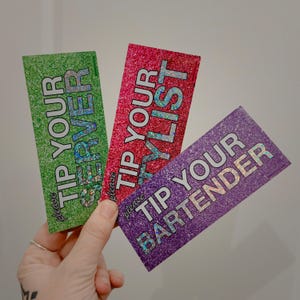 May include: Three glittery rectangular cards with the phrases "Tip Your Server", "Tip Your Stylist", and "Tip Your Bartender". The cards are green, red, and purple, with holographic text. The word "please" is at the bottom of each card.