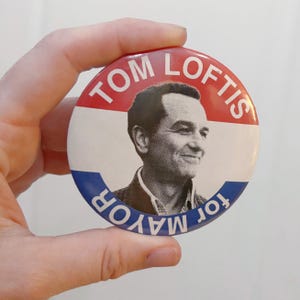 May include: A round political campaign button featuring a black and white halftone portrait of a man. The text TOM LOFTIS is printed in white on a red background at the top, and the text for MAYOR is printed in white on a blue background at the bottom.