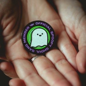 May include: A black and green enamel pin with a white ghost with a smiling face. The pin says "Official Ghoul Gang Spooky Member" in a purple font.