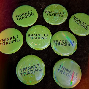 May include: A collection of eight circular lime green pin-back buttons featuring black text. Some buttons display the words TRINKET TRADING, while others display BRACELET TRADING. The surfaces have a holographic finish with various star and heart patterns.
