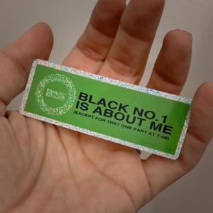 May include: A rectangular sticker with a green background and the text "BLACK NO.1 IS ABOUT ME (EXCEPT FOR THAT ONE PART AT 7:08)". The sticker has a glittery border and a circular design with the letter "O" in the upper left corner.