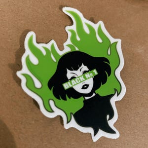 May include: A die-cut sticker featuring a stylized illustration of a woman with black hair and a black top, set against a green flame design. The text "BLACK No 1" is across the eyes. The sticker has a white border.