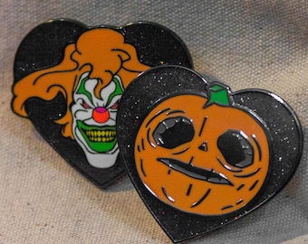 May include: Two heart-shaped enamel pins. One features a scary clown with orange hair, green and white face paint, and a red nose. The other pin is an orange jack-o'-lantern with a green stem. Both pins have a black glittery border.