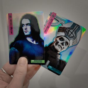 May include: Two holographic trading cards featuring portraits of musicians. One card shows Peter Steele with long black hair, the other depicts Papa Emeritus IV. Both cards have the text "HOTTIES of Metal" and are from the "OFFICIAL COLLECTOR CARDS" series.