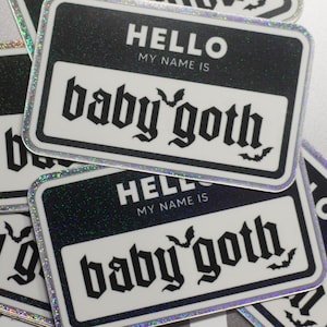 May include: A black and white name tag sticker with glitter trim. The sticker says "Hello, my name is baby goth" in a gothic font with bats on either side of the text.