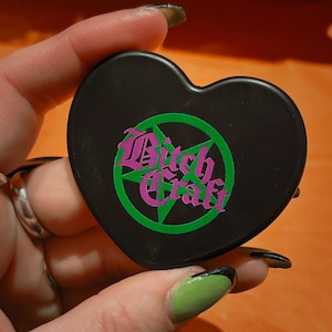 May include: Black heart-shaped compact with the words "Bitch Craft" in a gothic font, surrounded by a green pentagram. The text is in purple. The compact is held in a hand.