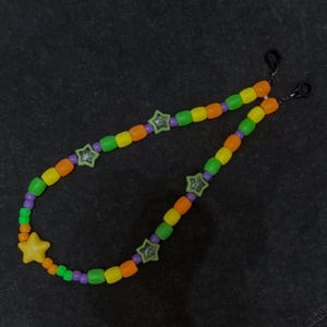 May include: A beaded eyewear chain featuring a repeating pattern of orange, yellow, green, and purple barrel-shaped beads. The chain is accented with several light green star-shaped beads and one large yellow star bead, finished with black rubber loops.
