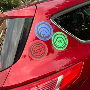 May include: A red car with three round stickers on the back. The stickers are black, green, and blue with white text and graphic designs. The text on the stickers reads "OT77M ROCKA PRATIC", "OT77M ROCKA PRATIC", and "OT77M ROCKA PRATIC".