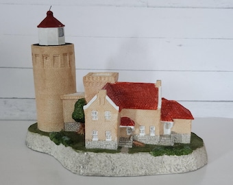 Puede incluir: Un detallado modelo de faro en miniatura con una torre de ladrillo beige, un tejado rojo y una luz blanca. El modelo incluye una casa con tejado rojo y pequeños árboles verdes, todo sobre una base de piedra.