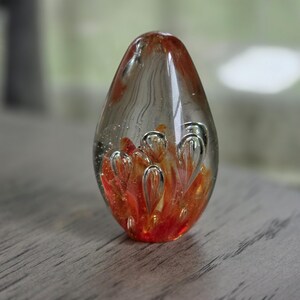 May include: A teardrop-shaped glass pendant featuring a clear exterior with an internal design of swirling orange and red hues. The glass contains numerous small air bubbles suspended throughout the interior, creating a textured, multi-dimensional appearance.