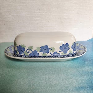 May include: White ceramic butter dish with a lid. The dish and lid are decorated with blue and green leaf patterns. The base has a blue and white patterned border. The dish is oval-shaped.