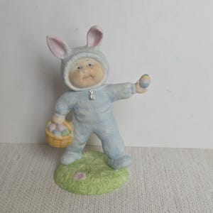 May include: A ceramic figurine of a child in a light blue bunny suit, holding a basket of Easter eggs and one egg. The figurine stands on a green base with a flower.