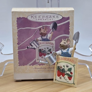 May include: A Keepsake Ornament box featuring a mouse figurine in overalls holding a watering can and a shovel. The box has a strawberry patch design with the word "STRAWBERRIES" and the text "For Decorative Use Only".