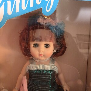 K&ouml;nnte beinhalten: Eine Vintage-Puppe mit rotbraunem Haar, die ein tealfarbenes Kleid und eine Perlenkette tr&auml;gt. Die Puppe hat gro&szlig;e Augen und tr&auml;gt einen blau-rosa Hut mit Schleier. Die Puppe befindet sich in einer Box mit dem Wort "Ginny" oben.