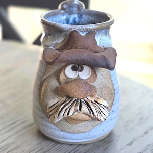 May include: A ceramic mug featuring a sculpted face of a person wearing a brown cowboy hat. The mug has a light blue and grey speckled glaze. The face includes large white eyes with dark pupils, a prominent nose, and a textured brown mustache.