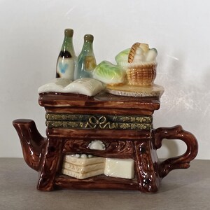 May include: A brown ceramic teapot shaped like a table, with a drawer and a handle. The top of the table has two bottles, a basket of food, and a book. The teapot is decorated with gold accents.