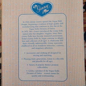 K&ouml;nnte beinhalten: Produktbox mit dem Ginny-Logo in Herzform in Blau. Der Text beschreibt die Geschichte der Ginny-Puppen, einschlie&szlig;lich ihrer Einf&uuml;hrung im Jahr 1951 und ihrer Anziehungskraft auf Sammler jeden Alters. Die Box hat einen hellrosa Hintergrund mit einem Herzmuster.
