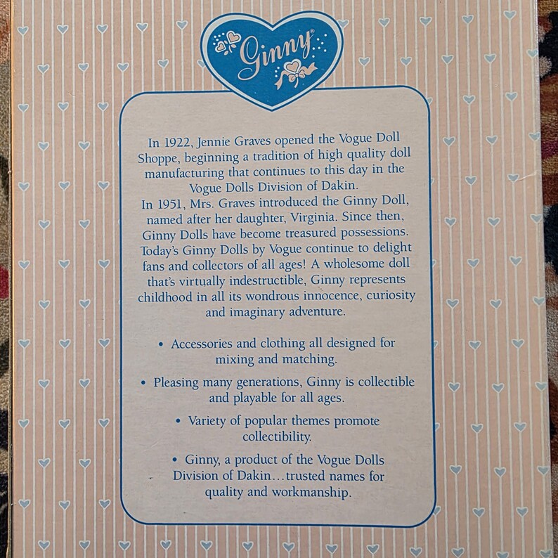 K&ouml;nnte beinhalten: Produktbox mit dem Ginny-Logo in Herzform in Blau. Der Text beschreibt die Geschichte der Ginny-Puppen, einschlie&szlig;lich ihrer Einf&uuml;hrung im Jahr 1951 und ihrer Anziehungskraft auf Sammler jeden Alters. Die Box hat einen hellrosa Hintergrund mit einem Herzmuster.