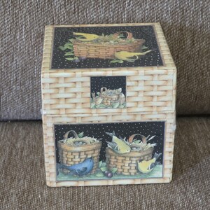 May include: A decorative box with a woven basket design. The box features illustrations of birds, nests, and baskets filled with greenery and berries. The background is black with white polka dots. The box is beige and square.