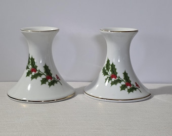May include: Two white ceramic candle holders with a gold rim and a holly berry design. The candle holders have a flared base and a narrow top. The holly design features green leaves and red berries.